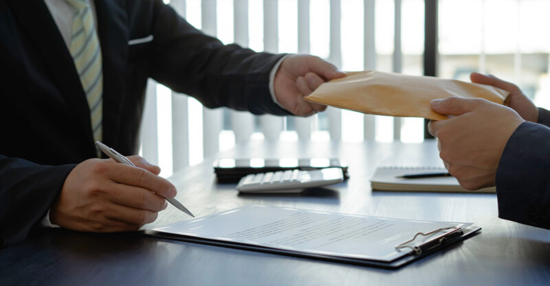 Men in suits with contract paper on desk, one man handing the other a thick envelope.