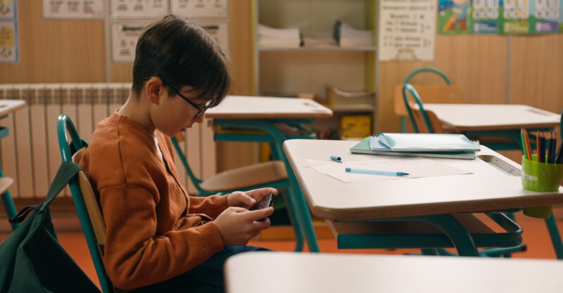 Schoolboy playing video game on mobile phone at lesson at school and hiding smartphone under class desk.