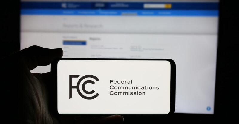 Person holding a smartphone with FCC logo and computer in background on FCC website.