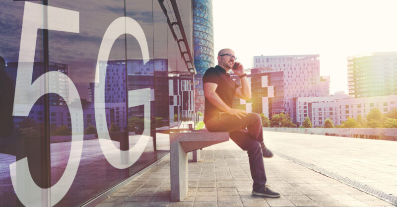 Man using a smartphone with the new 5G mobile network sitting on a bench in front of a building with large "5G" sign on window.