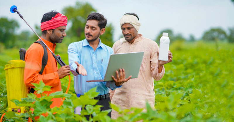 Young indian agronomist giving liquid fertilizer bottle to farmer and showing product information in laptop at green agriculture field.