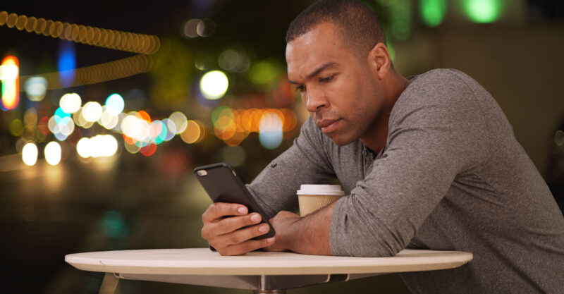 Man sitting alone at outdoor cafe drinking coffee at night and browsing the internet on smartphone.