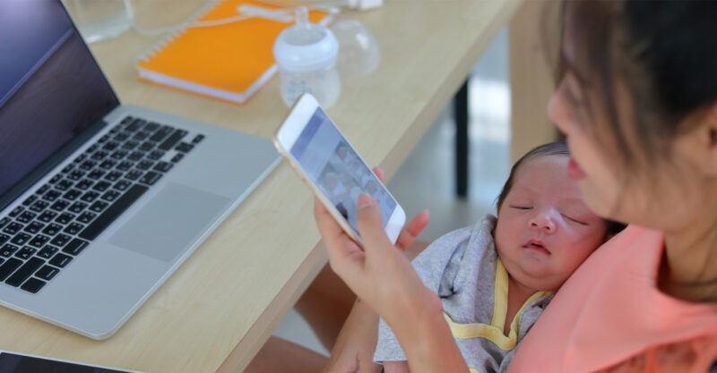 Mother holding cell phone and infant next to a laptop.