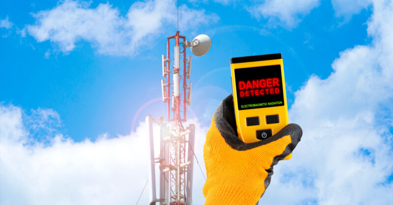 Hand with glove on holding an EMF meter up in front of a cell tower.