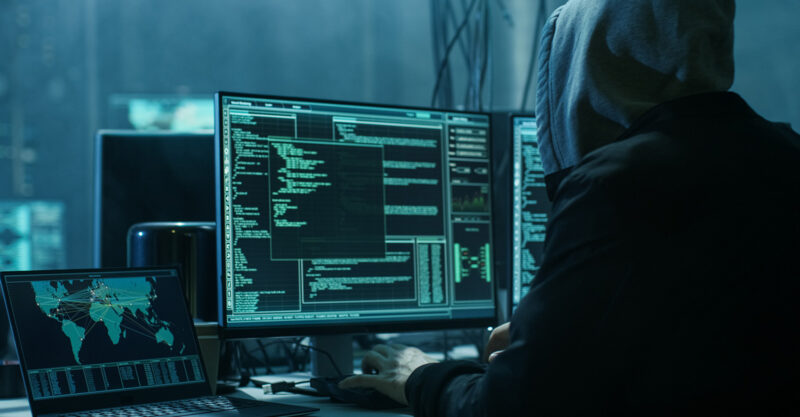Dangerous Hooded Hacker in His Hideout Place has Dark Atmosphere, Multiple Displays, Cables Everywhere.