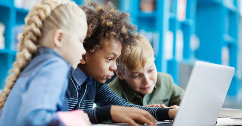 Group of three excited diverse students using laptop sitting at desk in school